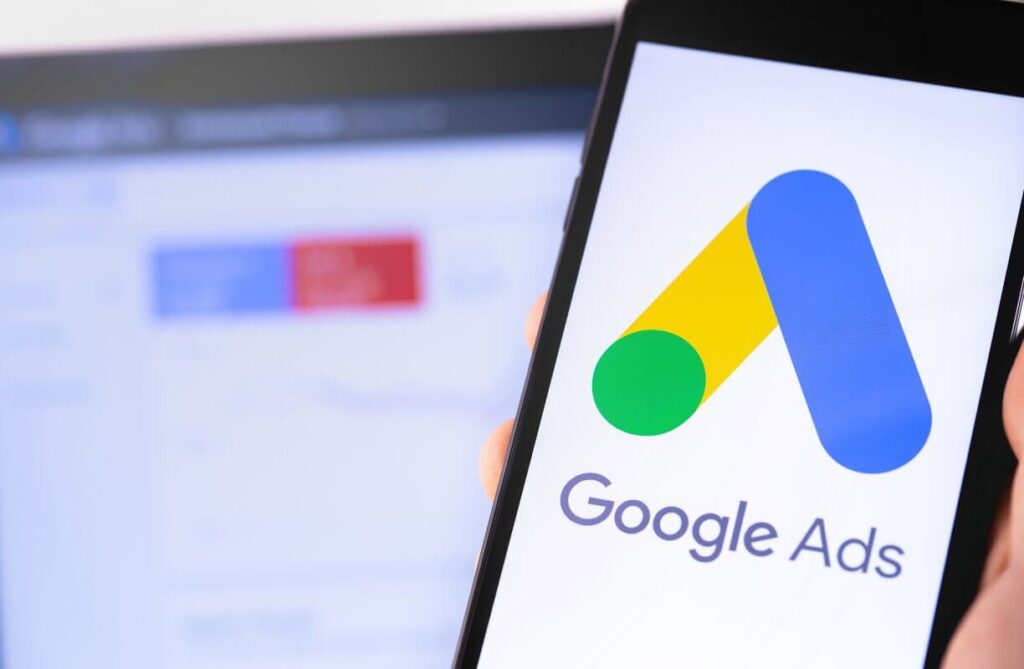 un telephone affichant le logo de google ads avec en fond l'écran d'un ordinateur